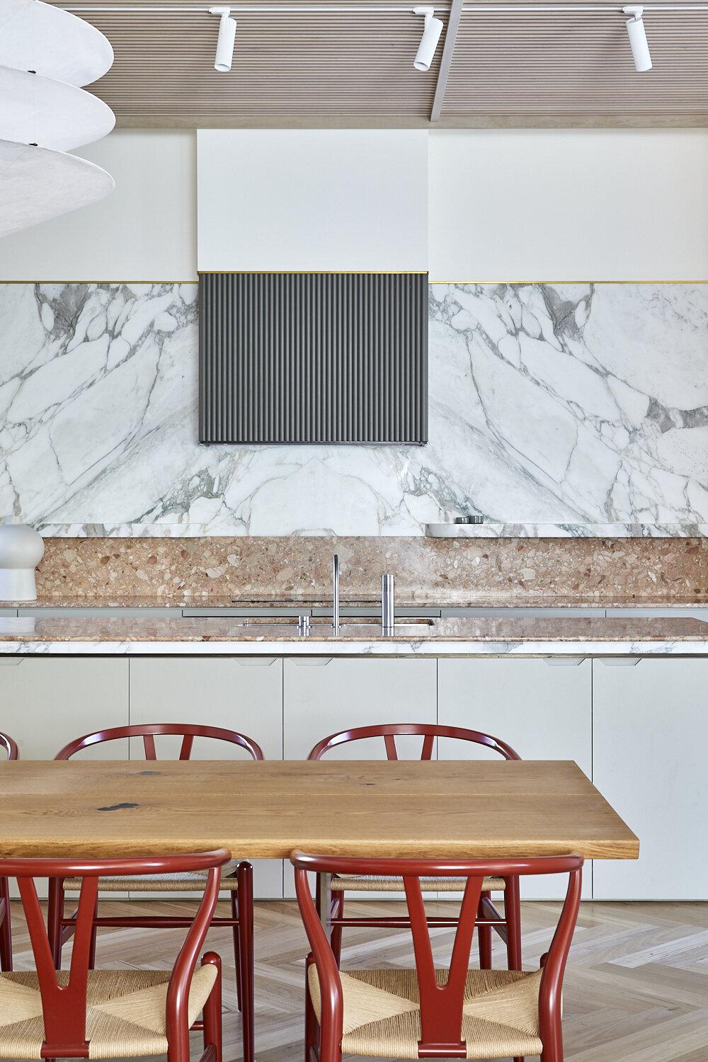 Contemporary kitchen with a wooden dining table, red chairs, marble backsplash, and minimalist decor, creating a clean, modern aesthetic.