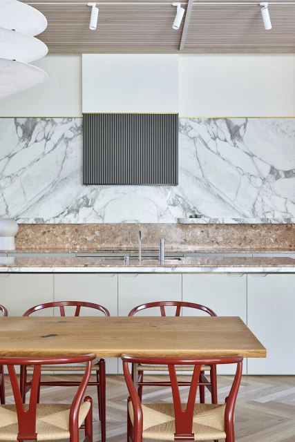 Contemporary kitchen with a wooden dining table, red chairs, marble backsplash, and minimalist decor, creating a clean, modern aesthetic.