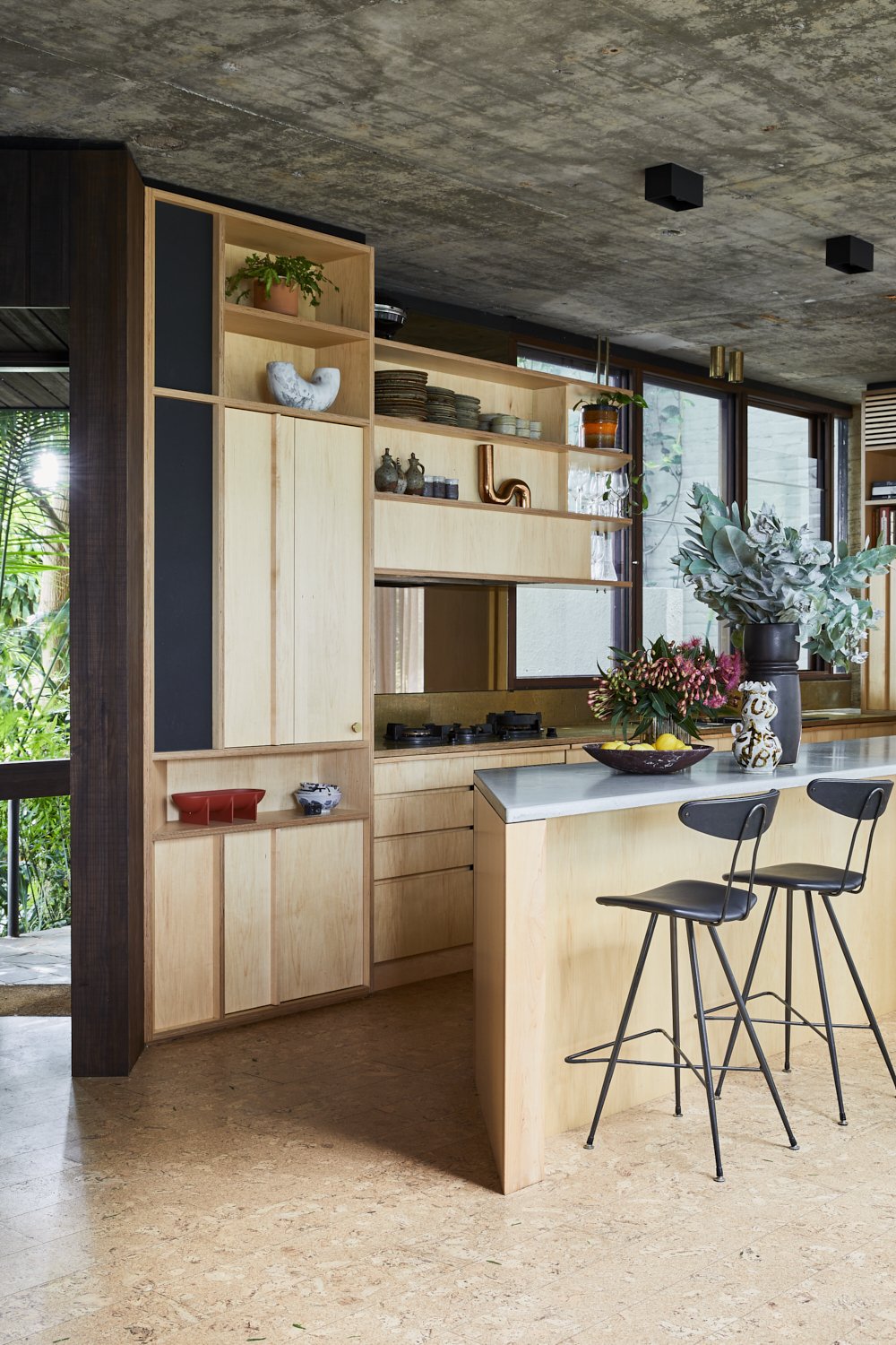Interior shot of a modern kitchen featuring light wood cabinetry, a grey countertop, and two bar stools, illuminated by natural light.