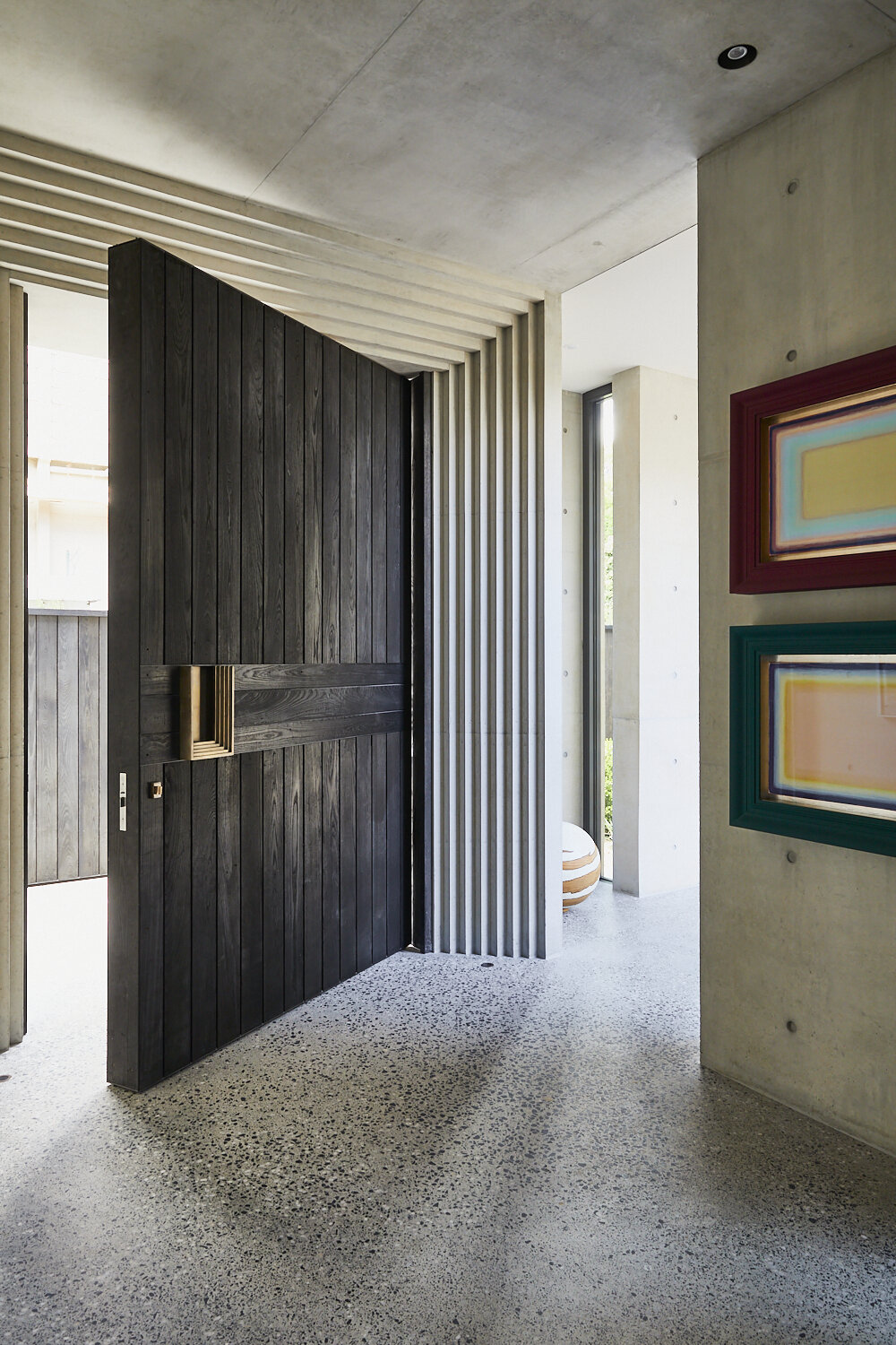 Dark wood pivot door open in a modern, minimalist foyer with concrete walls, grey terrazzo floor, and framed artwork.