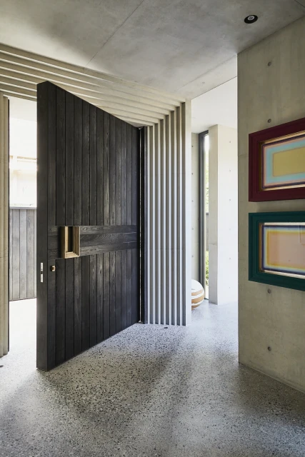 Dark wood pivot door open in a modern, minimalist foyer with concrete walls, grey terrazzo floor, and framed artwork.