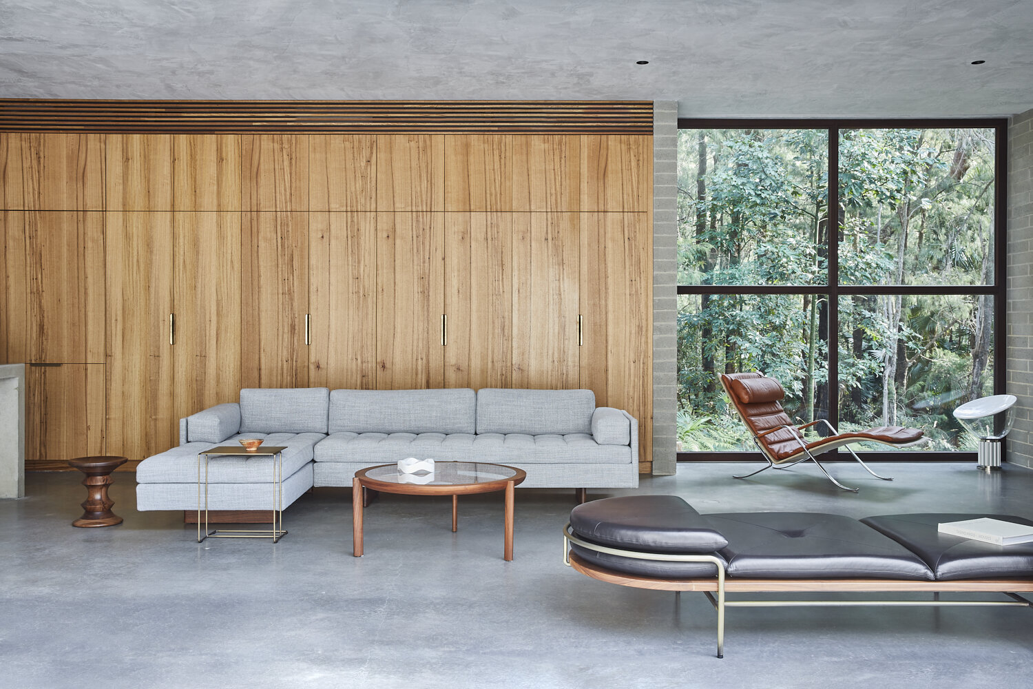 Modern living room featuring a grey sectional sofa, wood wall paneling, lounge chairs, glass-top coffee table, and large window with forest view.