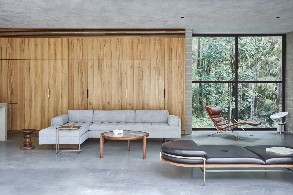 Modern living room featuring a grey sectional sofa, wood wall paneling, lounge chairs, glass-top coffee table, and large window with forest view.
