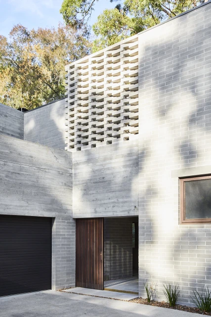 Exterior of a modern grey brick home featuring a unique breeze block wall, a dark garage door, and a slatted wood front door.
