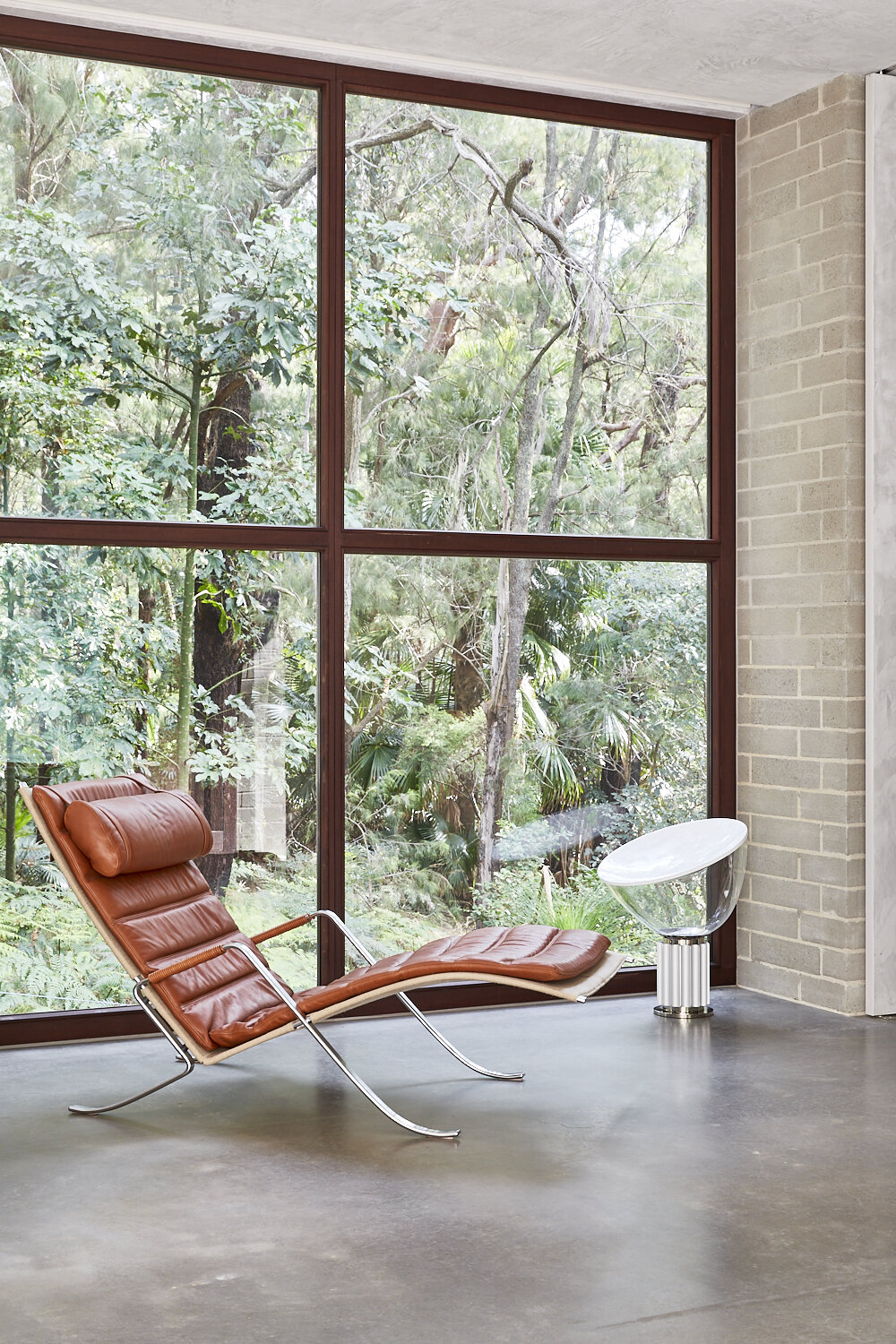 Tan leather chaise lounge faces a window with green forest background. Modern architectural interior with gray floor and natural light.