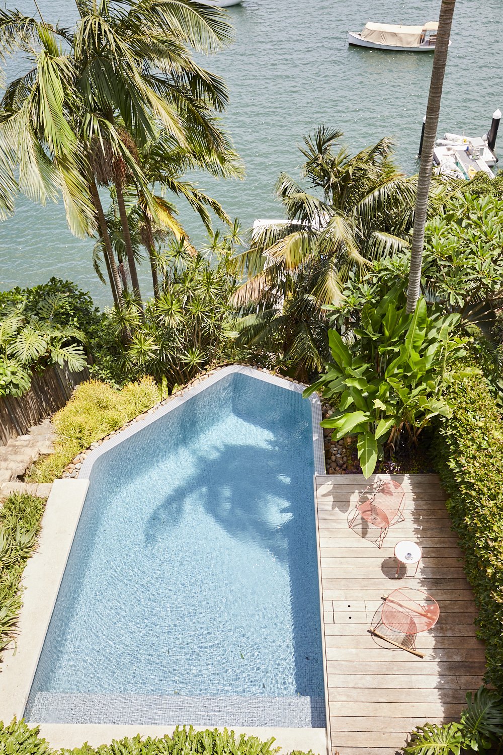 Aerial view of a rectangular swimming pool with blue water, surrounded by a wooden deck, lush tropical plants, and ocean in the background.