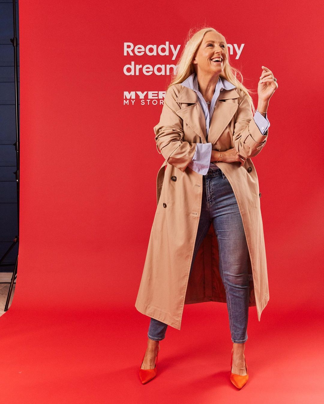 A blonde woman smiles wearing a trench coat, blouse, jeans, and pointed-toe heels, posed in a full shot in front of a red backdrop.