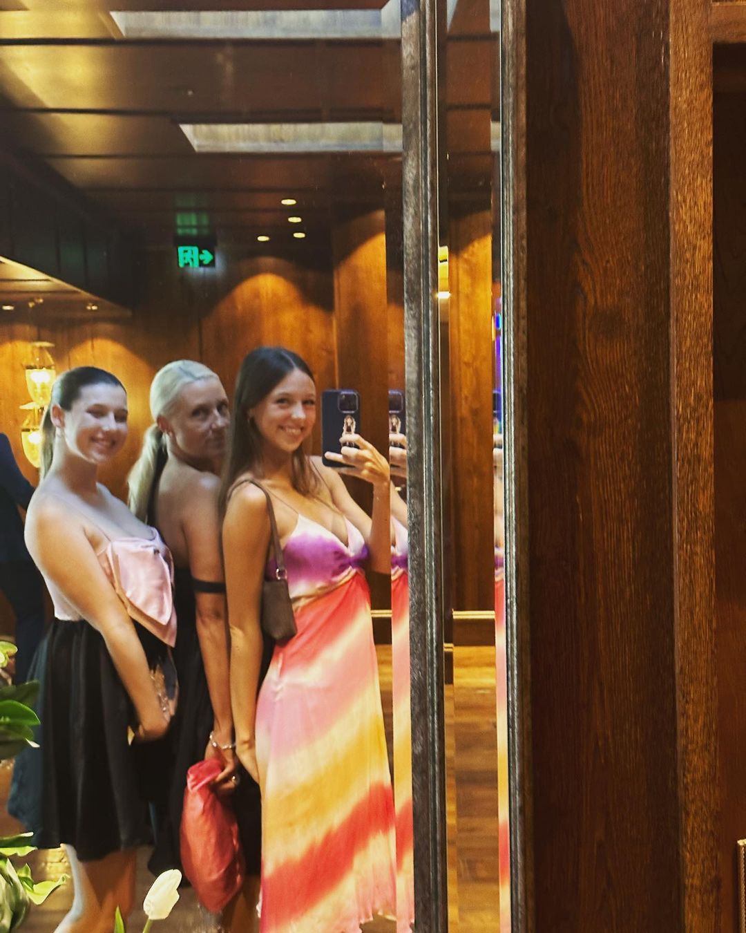 Three women pose in front of a mirror in an indoor environment with wood paneling, dresses, and cell phone visible.