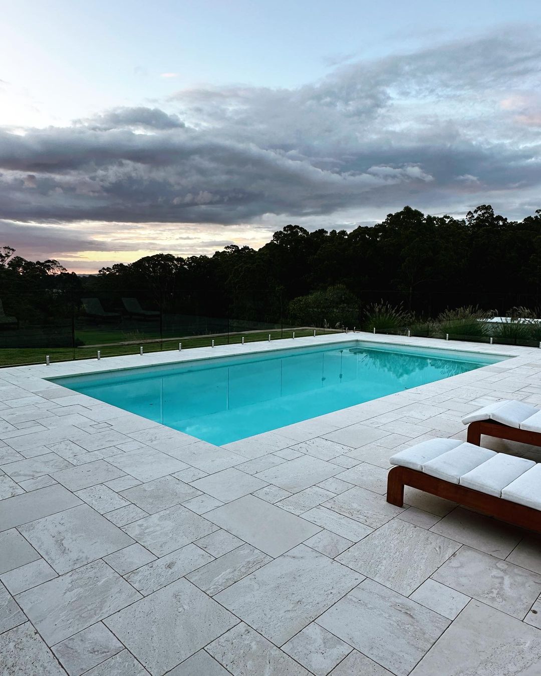 Outdoor swimming pool with light-blue water and travertine tile surround, next to lounge chairs, with dark green trees and cloudy sky.