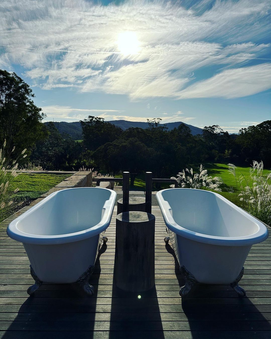 Outdoor bathroom with two clawfoot tubs on a wooden deck, overlooking a green field and forested hills under a bright, partly cloudy sky.