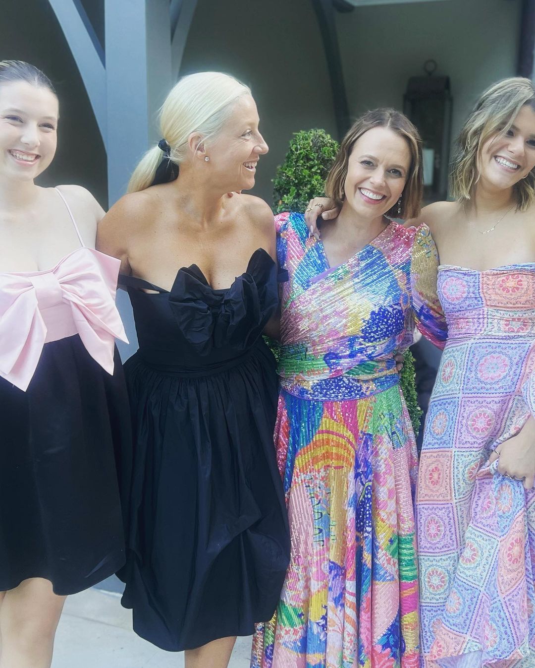 Four women pose together outdoors. They're wearing colorful dresses and are smiling, with a building visible in the background.