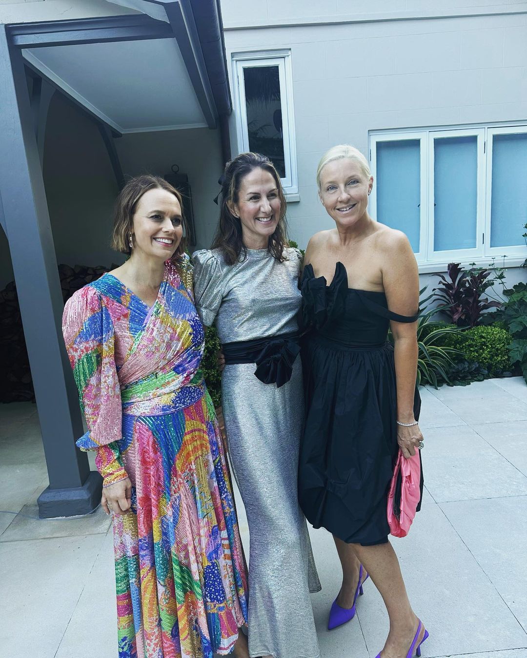 Three women pose in colorful dresses outside a building with garden foliage. Silver, black, and print designs showcase fashion details.