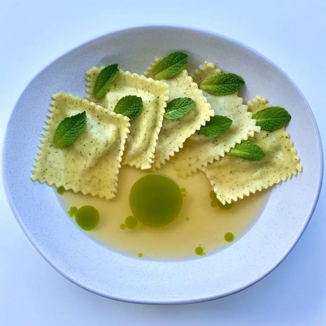 Overhead shot of ravioli pasta with mint leaves in a white bowl, complemented by small droplets of green oil on a white backdrop.