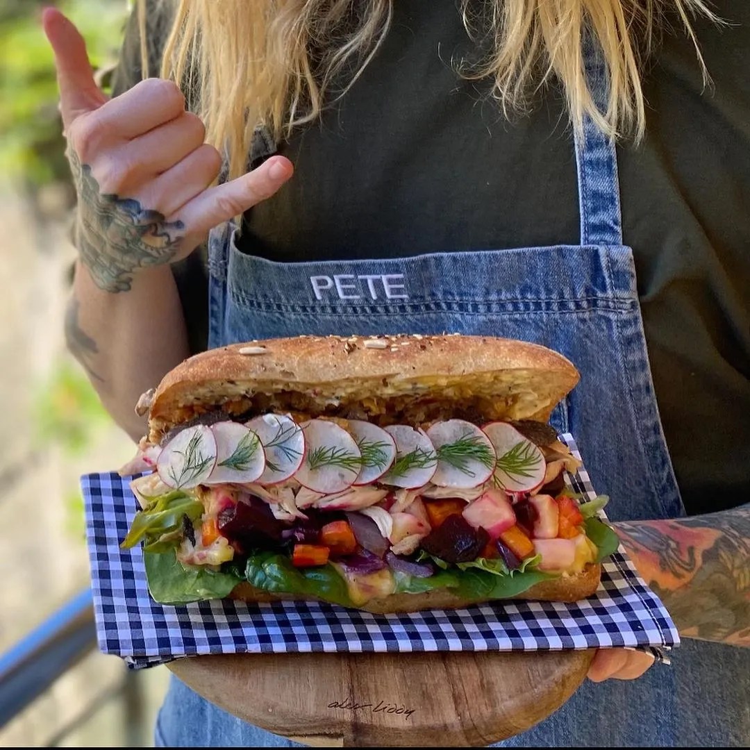 A large sandwich filled with radish, beet, and greens is presented on a checkered napkin and wooden board, held by a person.
