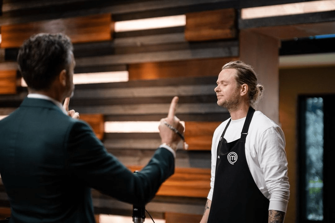 Two men in a cooking studio. One man in a suit gestures with a raised finger to another chef wearing an apron.