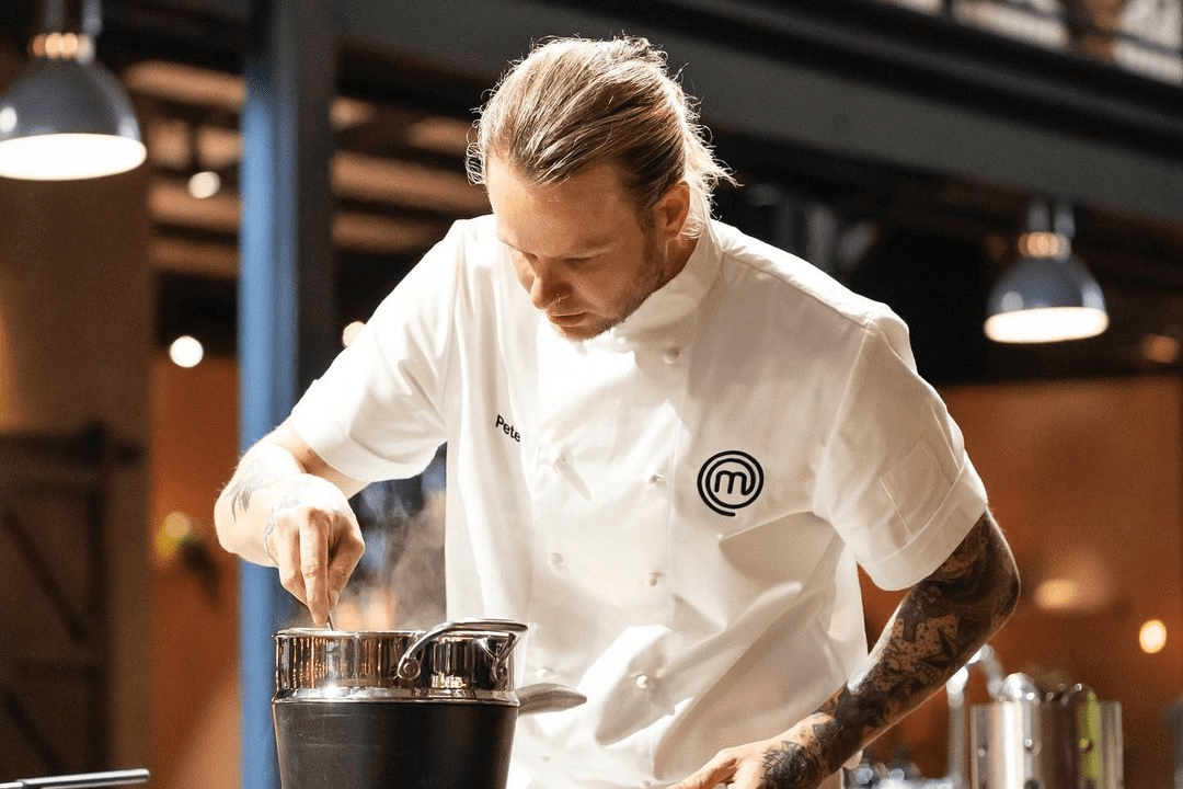 Male chef in a white chef's coat is stirring ingredients in a metal pot in a professional kitchen, illuminated by overhead lights.