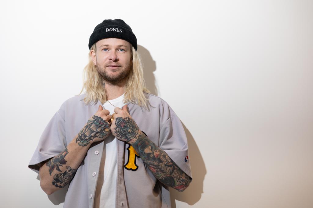 Man with long blond hair and tattoos wearing a baseball jersey and beanie against a white wall in a studio setting.