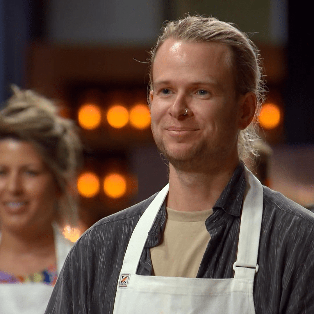 Smiling blond man in chef's whites stands indoors with background lights. Cooking, food preparation, studio lighting.