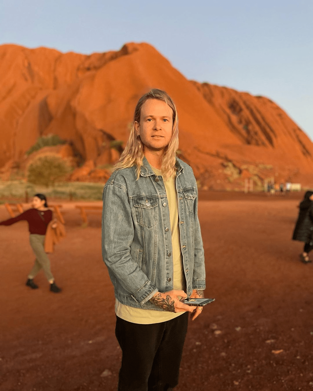 Blonde man poses with a mobile phone in front of Uluru, Australia, wearing a denim jacket, with other people and red desert sand.