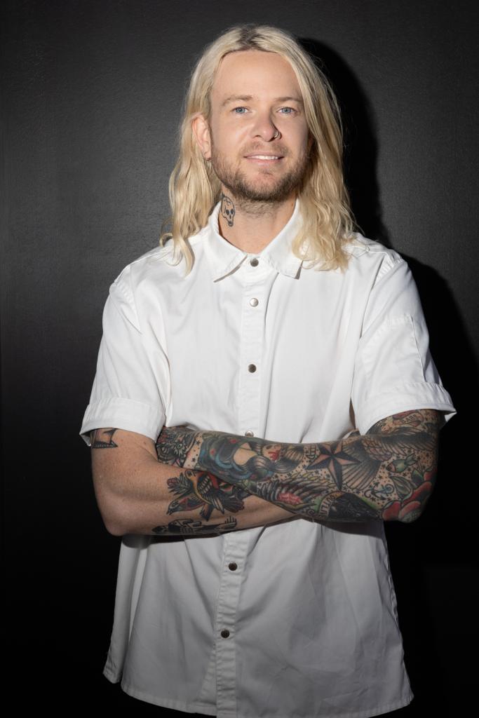 Portrait of a man with long blonde hair and extensive tattoos wearing a white button-down shirt against a dark grey backdrop.