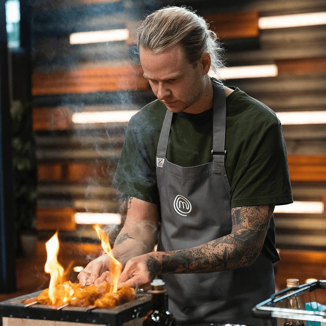 Man in apron cooking food on open flame grill indoors, culinary arts and food preparation in a cooking show environment.