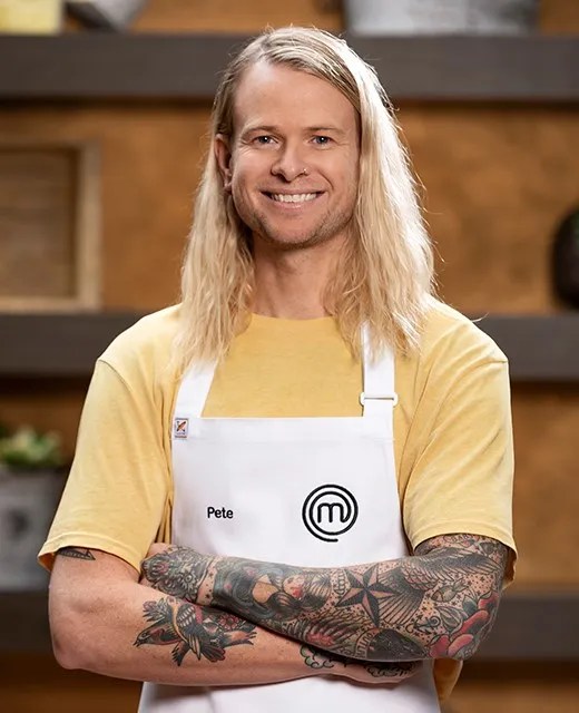 Smiling man with long blond hair, wearing a yellow shirt and white apron with the MasterChef logo, stands indoors with tattooed arms crossed.