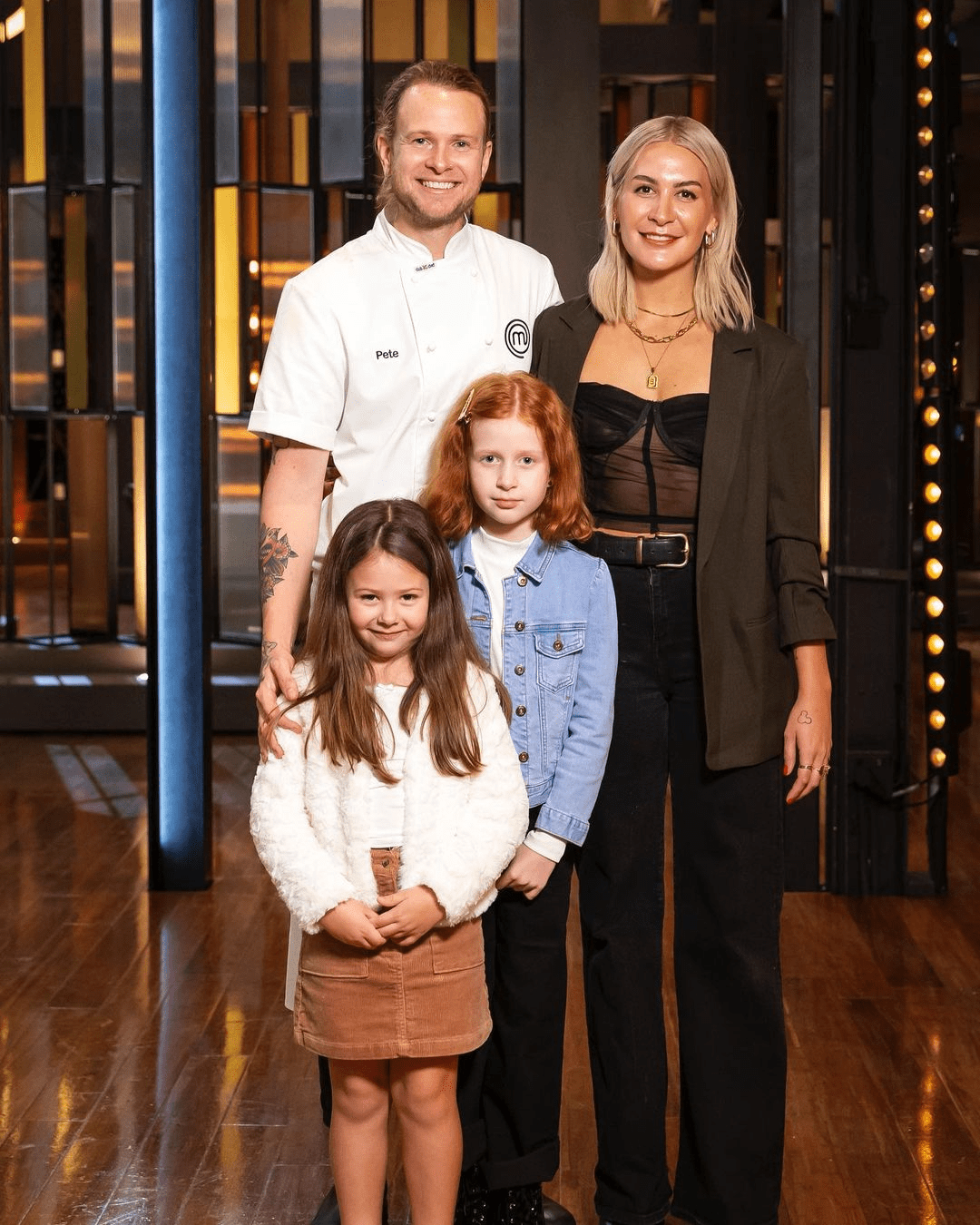 Family portrait of two adults and two children posing indoors with a studio backdrop and wood floor, family members are smiling.