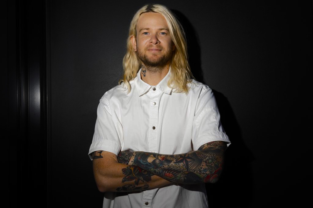 Portrait of a blonde adult male with tattoos on his arms, standing with crossed arms against a dark background. He wears a white button-down shirt.