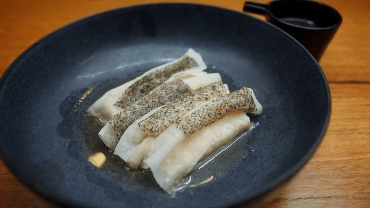Close-up of white fish fillets arranged on a dark plate, complemented by a small black cup, set on a wooden table for a simple, elegant food presentation.