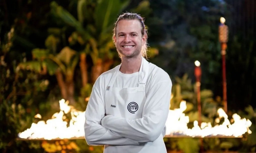Portrait of smiling adult male wearing a white chef's coat and apron, arms crossed, standing in a garden at night near flaming torches.