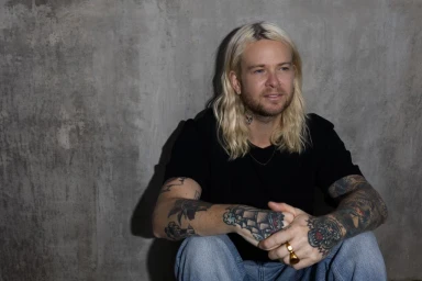 Blonde man with tattoos wearing a black t-shirt and blue jeans seated against a textured concrete wall.