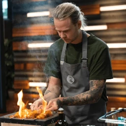 A man wearing a grey apron over a green t-shirt cooking over an open flame on a small grill in a kitchen.