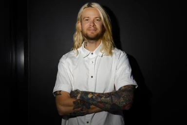 Portrait of a man with long blonde hair and tattoos wearing a white button-down shirt against a dark background