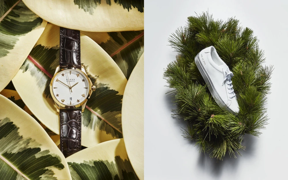 Elevated shot of an analogue Gucci watch on leaves and a white sneaker surrounded by a pine wreath on a white background.