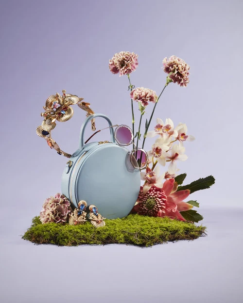 Fashion still life: light blue purse surrounded by pink and white flowers, green moss, jewelry, and sunglasses against a light purple backdrop.
