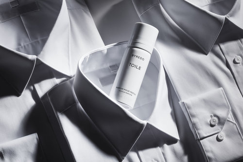 Close-up of a white perfume bottle, Byredo Toile, resting on a pile of white cotton dress shirts creating a minimalist, clean aesthetic.