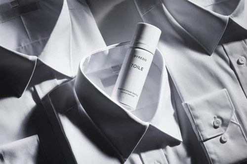 Close-up of a white perfume bottle, Byredo Toile, resting on a pile of white cotton dress shirts creating a minimalist, clean aesthetic.