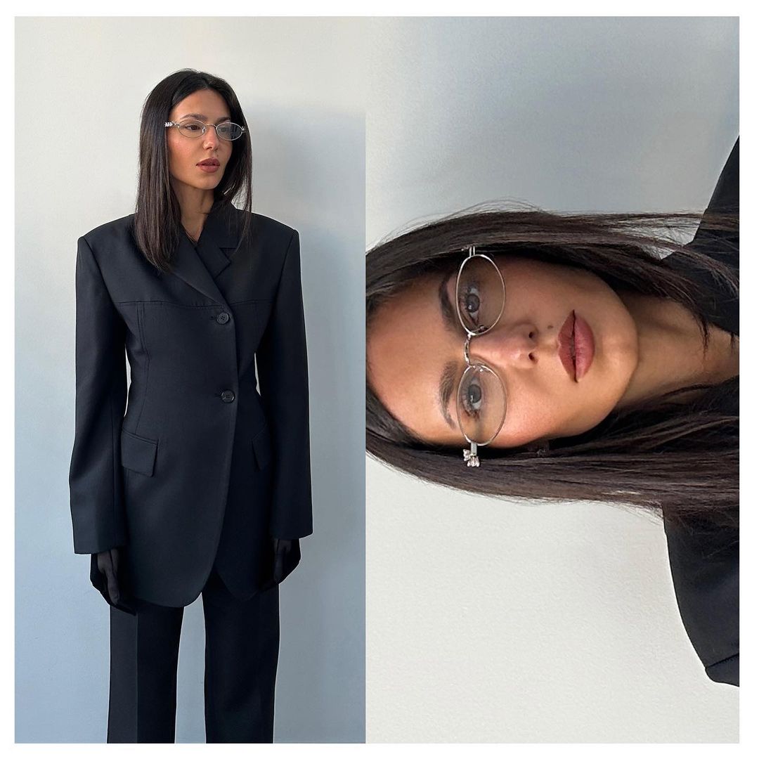 A young woman poses in a fashionable black suit and glasses against a white wall, creating a studio-style portrait.