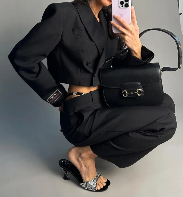 A woman poses in a black crop jacket and pants, with a black purse and bedazzled high heel in a studio environment.