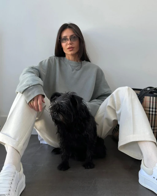 Brunette woman in a grey sweatshirt poses with a black Griffon dog, sitting on the floor in front of a white wall.