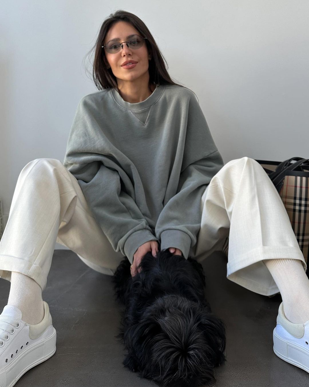 Woman in sweatshirt sitting with dog indoors with Burberry tote bag in background; casual fashion, simple background, natural light.