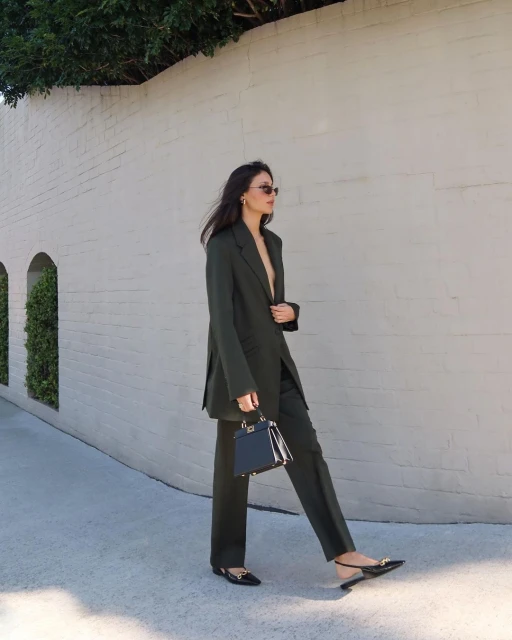 Adult woman walking in an olive green pantsuit, holding a small black handbag, with a light-colored brick wall in the background.
