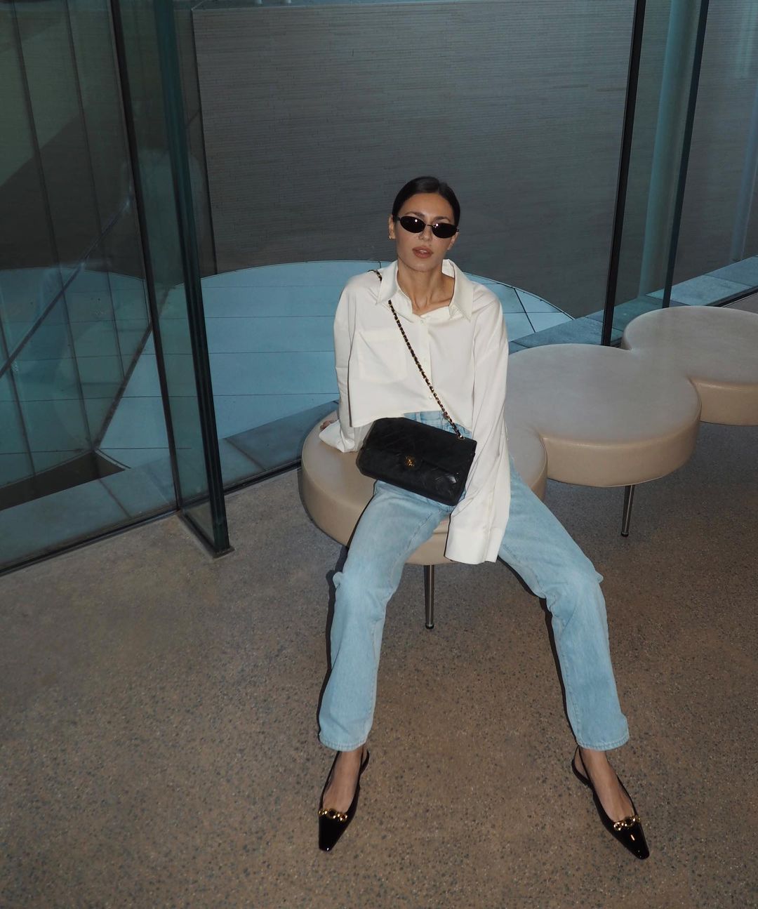 A woman with dark hair and sunglasses sits on a stool, wearing a white button-down shirt, light blue jeans, black flats, and a black purse.