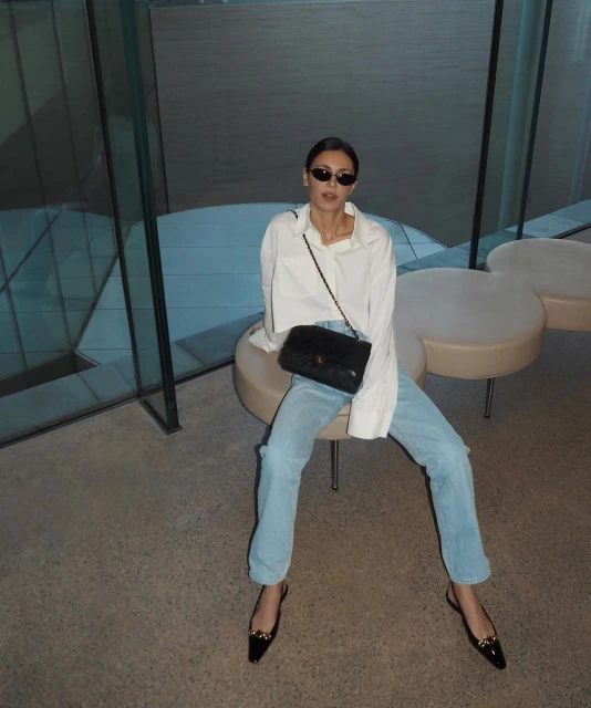 A woman with dark hair and sunglasses sits on a stool, wearing a white button-down shirt, light blue jeans, black flats, and a black purse.