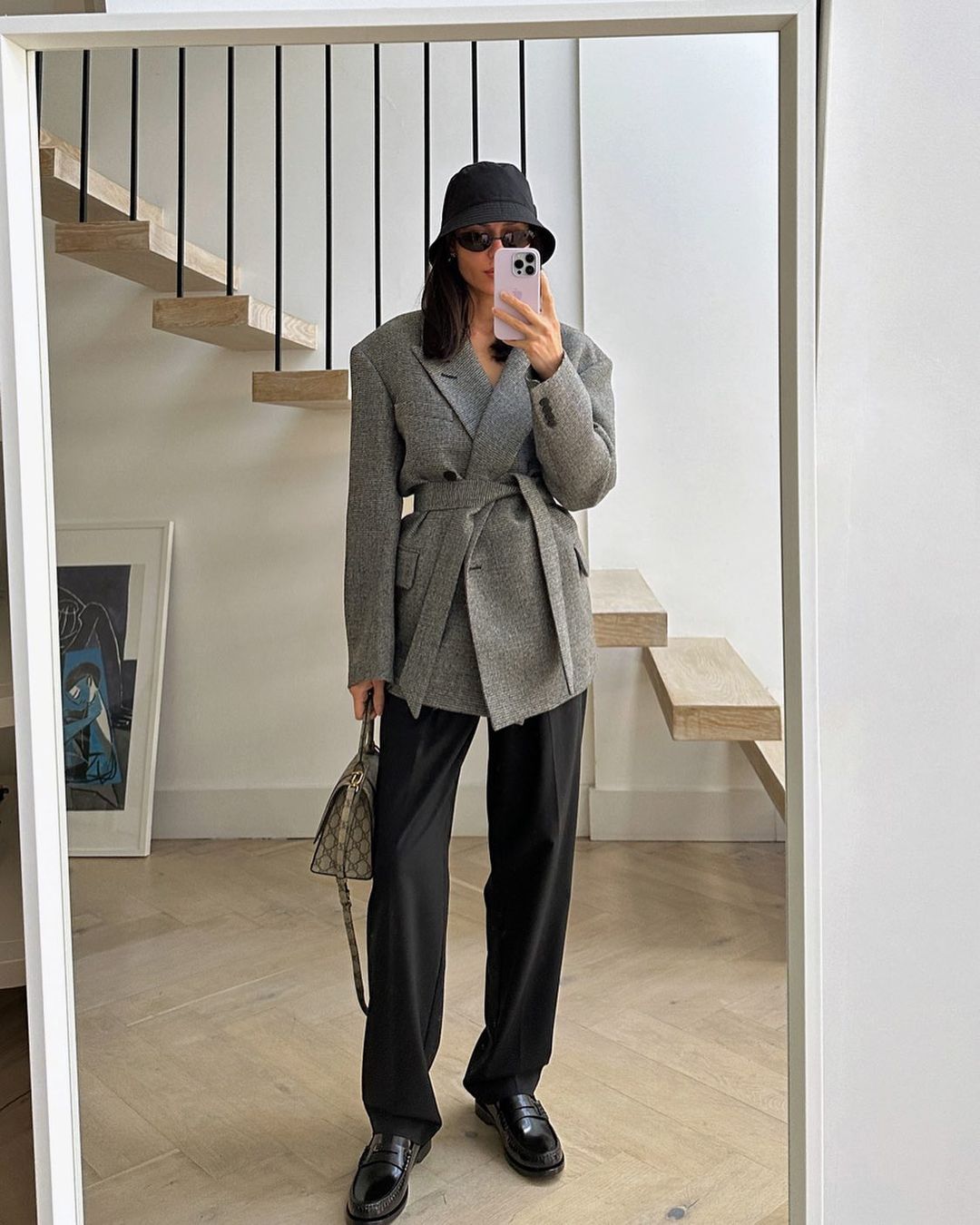 Woman in oversized grey blazer and black bucket hat takes mirror selfie in bright indoor setting, showcasing fashion and personal style.