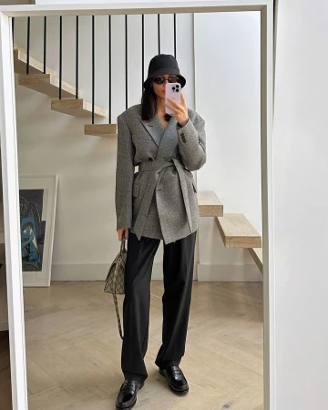 Woman in oversized grey blazer and black bucket hat takes mirror selfie in bright indoor setting, showcasing fashion and personal style.