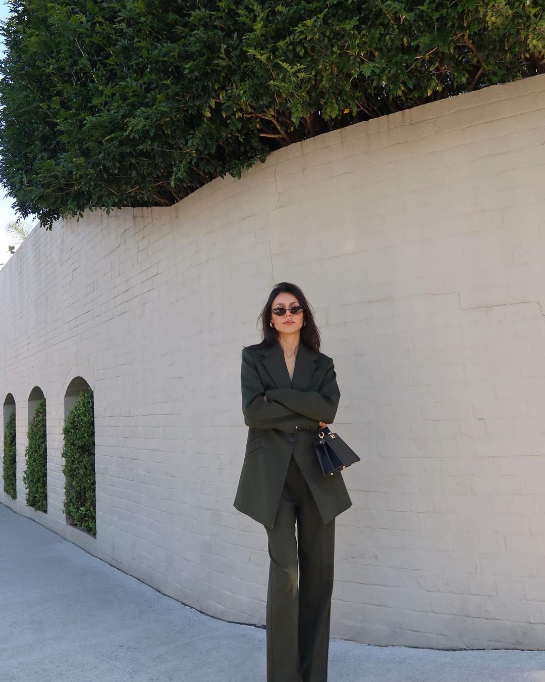 Adult female in an olive-green pant suit stands against a white wall with green hedges, holding a small dark purse with folded arms.
