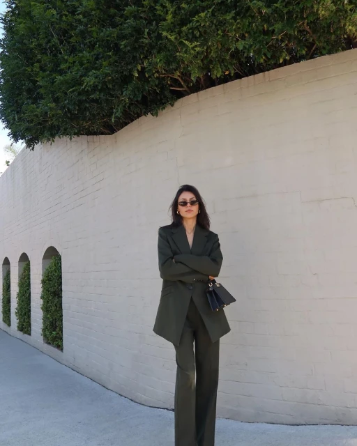 Adult female in an olive-green pant suit stands against a white wall with green hedges, holding a small dark purse with folded arms.