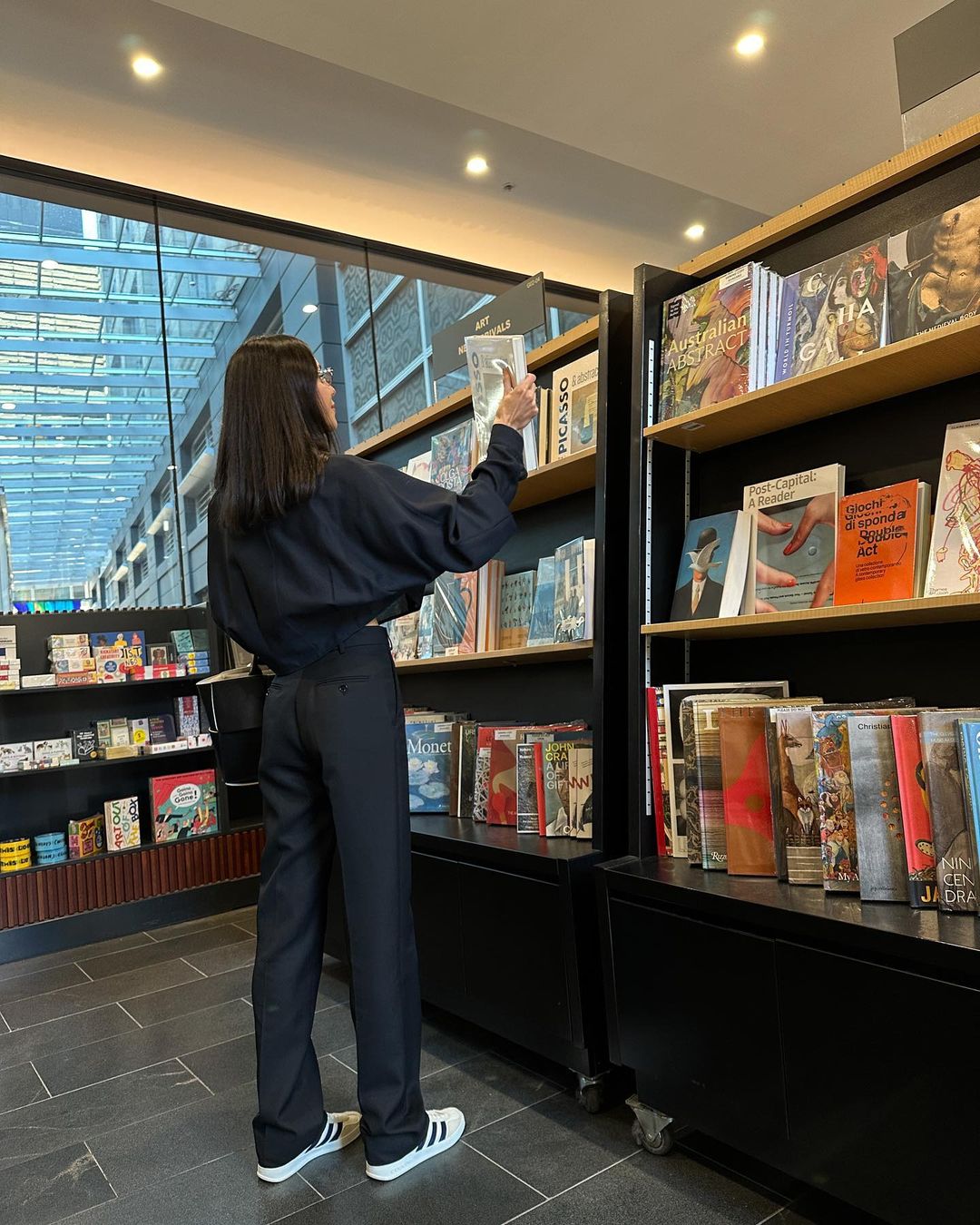 Woman in black outfit reaches for a book in a bright bookstore, surrounded by shelves filled with books, on a modern tiled floor.