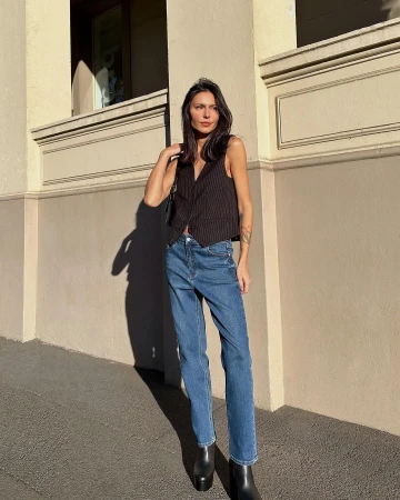 Woman wearing a dark pinstripe vest and blue jeans standing against a cream building exterior in Sydney